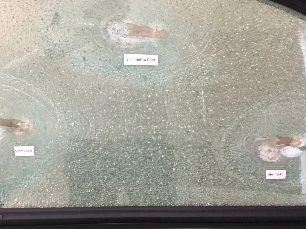 A vehicle window after a 9mm test: the glass is crazed but the rounds are stopped, with labels reading 9mm Solid and 9mm Hollow Point