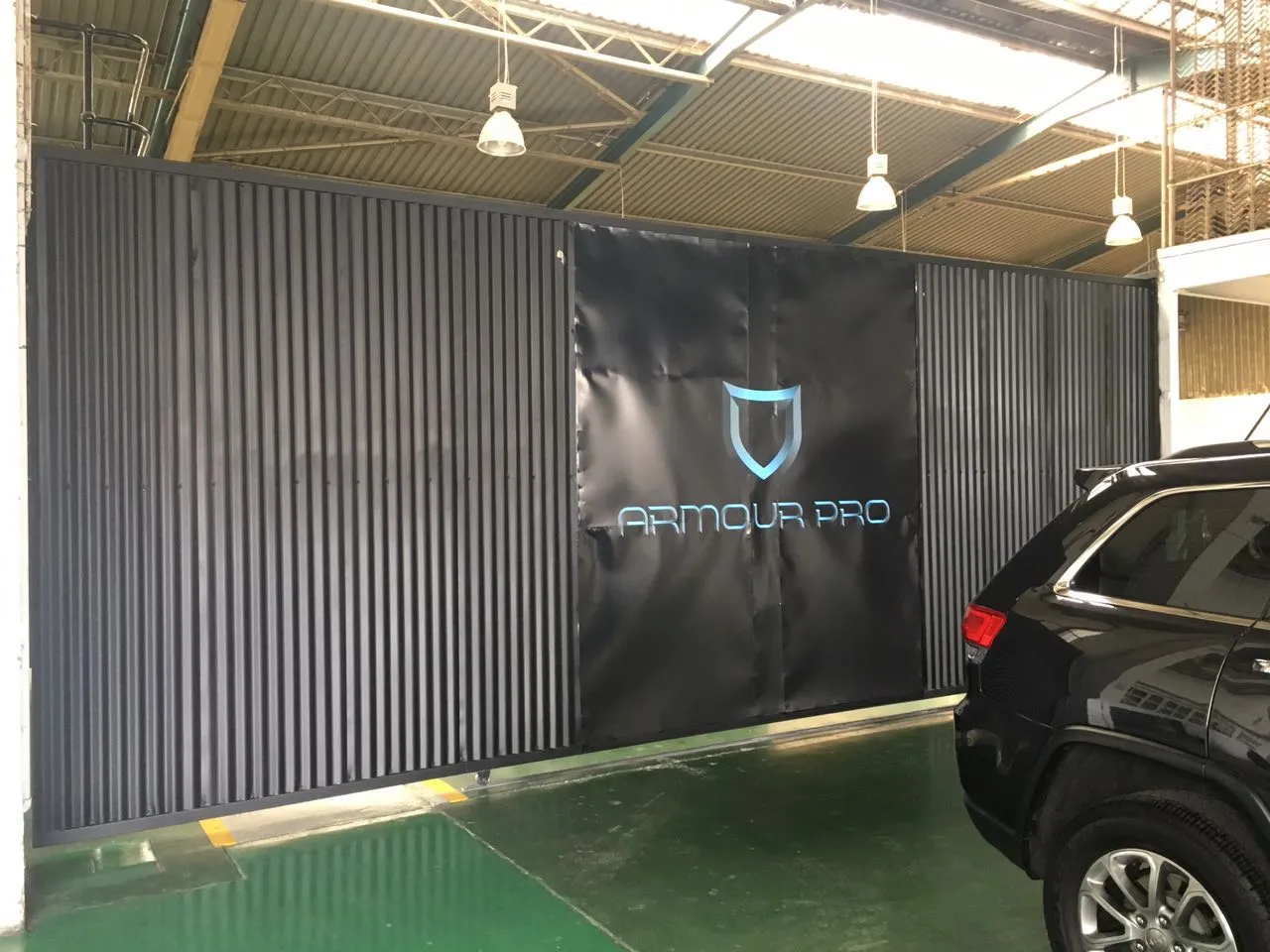 Inside the Armour Pro workshop: a black wall banner with the Armour Pro shield logo, with a black SUV parked on the green floor beside it